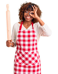 Poster - Young african american woman wearing apron holding rolling pin smiling happy doing ok sign with hand on eye looking through fingers