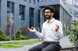 © Liubomir - Smiling and relaxed young Indian male businessman sitting on bench in lotus position near offices, meditating and resting with eyes closed.