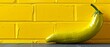   A green banana rests on a table beside a yellow brick wall and in front of a yellow wall