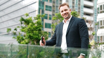 Skilled caucasian business man looking at camera while standing at rooftop. Professional investor smiling with confident while wearing formal suit at modern building in green city. Lifestyle. Urbane