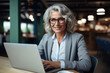 A poised senior businesswoman with sophisticated silver curls confidently uses her laptop in a bright, modern office environment, her professional attire speaking to her experience
