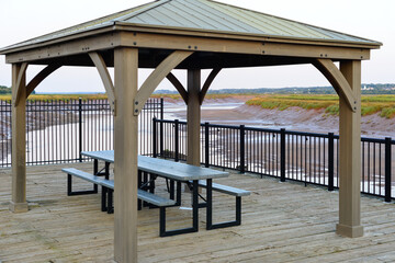 A large four-sided wooden gazebo overlooking the Bay of Fundy.  The roof is covered in green metal panels. There's a green picnic table under the arbor. The river is low with red mud on both sides.