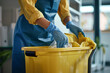 gloved hands of female cleaner throwing trash from garbage bin into plastic bucket on janitor trolley