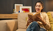 © Chanakon - Asian beautiful woman sitting reading book on cozey couch sofa in living room with warm light at night. People female stay home part time relax at home.