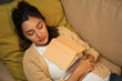 © Chanakon - Sleeping Asian beautiful woman lay down on cozey couch sofa while reading book in living room with warm light at night. People female reading book until fall asleep stay home part time relax at home