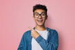 Excited happy black curly haired man in braces, open mouth, wear glasses denim shirt advertise show peek point area for sales slogan text, isolated rose pink background. Dental care ophthalmology ad.