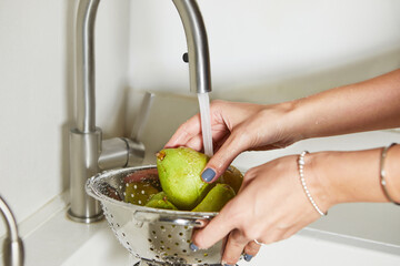 Woman in white shirt washing green pears under clear water in stainless steel kitchen sink