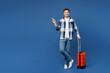 Traveler man wears shirt casual clothes hold suitcase bag point finger aside isolated on plain blue background. Tourist travel abroad in free spare time rest getaway Air flight trip journey concept.