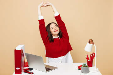 Wall Mural - Young relaxed employee business woman she wear red sweater shirt sit work at office desk with pc laptop stretch hands have break isolated on plain pastel beige background. Achievement career concept.