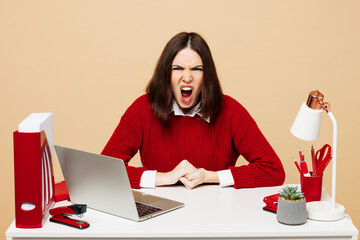 Wall Mural - Young sad mad angry outraged employee business woman wear red sweater shirt sit work at office desk with pc laptop scream shout isolated on plain pastel beige background. Achievement career concept.