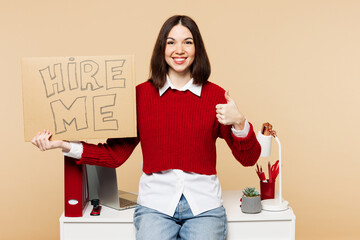 Wall Mural - Young employee business woman wear red sweater shirt work at office sit on desk with pc laptop hold cardboard sign card hire me isolated on plain pastel beige background. Achievement career concept.