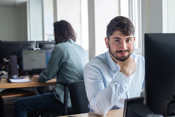 Poster - portrait d'un jeune homme employé de bureau souriant quoi est assis à son poste de travail devant son ordinateur. Un collègue est assis derrière lui.