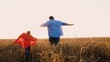 Father and son superhero in red cloak flying running at sunny dry wheat field back view. Happy family parent and son playing imagination powerful brave hero fantasy character costume rural outdoor