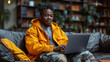 Young man in yellow jacket using laptop on couch with bookshelves in background.