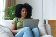 Young African American woman wearing jeans and green t-shirt sitting working in laptop in stylish white home