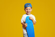 Charming blonde boy holding blue longboard and smiling while looking at camera. Isolated over yellow background.