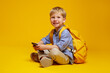 Joyful pupil boy in blue shirt with yellow backpack holding mobile phone in hands, smiling at camera while sitting on floor with crossed legs, isolated over orange background.