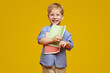 Excited little nerd kid in blue shirt smiling and holding two notebooks while being ready for school, isolated against yellow background.