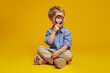 Positive curious schoolboy sitting on studio floor with crossed legs while looking away through magnifying glass, isolated over yellow background.