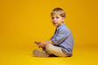 Horizontal photo of little handsome kid with blonde hair in casual clothes looking away while sitting with crossed legs on studio floor, holding smartphone, isolated over yellow background