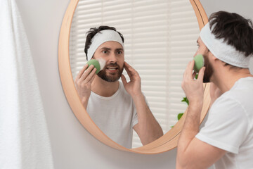 Sticker - Emotional man with headband washing his face using sponge near mirror in bathroom