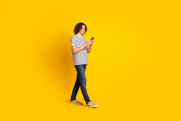 Poster - Full size portrait of nice young man walk empty space hold phone wear striped t-shirt isolated on yellow color background