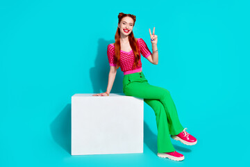 Wall Mural - Full length photo of excited friendly lady dressed pink knitted shirt sitting cube showing v-sign empty space isolated blue color background