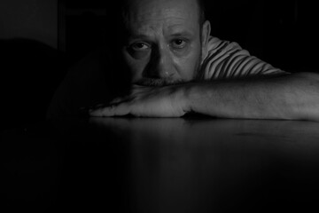 Wall Mural - Grayscale closeup of a man in the darkness leaning on the table and looking at the camera.