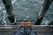 A contemplative scene with a man's feet on a wooden pier over serene water, bordered by weathered pillars