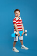 Vertical photo of full body little kid boy with headphones on neck, wearing trendy outfit, holding skateboard while standing isolated on blue backdrop. Childhood lifestyle concept