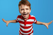 Close up portrait of cheerful little kid boy in red striped polo shirt celebrating with raised arms isolated over blue background. Children studio portrait.