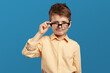 Little sad boy with curiosity looking through glasses at camera on blue backdrop. Doubtful kid wearing beige shirt and staring with suspicion at camera.