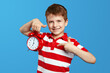 Excited little kid boy in casual red striped shirt, pointing index finger on red alarm clock. Posing isolated on blue background studio portrait. People emotions lifestyle concept. Mock up copy space.