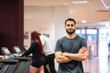© JoseIMartin - Casual man standing confidently with arms crossed at the gym, others exercising behind.