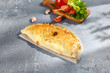 Golden calzone pizza on parchment paper, with fresh tomatoes and garlic in the background on a textured grey surface
