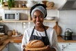A smiling young black woman in an apron stands in a bright kitchen and holds a wooden baking board on which lies freshly baked bread
