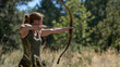 An athletic woman in a forest setting draws a longbow with precision, her posture poised in the midst of vibrant greenery