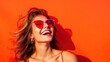 A joyful young woman wearing sunglasses, laughing and enjoying the sunshine, against a vivid orange background