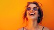 A joyful young woman wearing sunglasses, laughing and enjoying the sunshine, against a vivid orange background