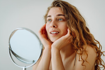 Wall Mural - Young woman with red spot unhappy about acne and skin imperfections, looking at reflection in mirror in bathroom. Facial skin care. Acne problem.