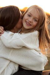 Wall Mural - Family day, mother's day. Happy smiling young mom and adorable child daughter soft hugging, kissing and spending time together at autumn on sunset. Idyllic family having fun outdoors on fall holiday.