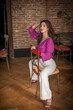 Beautiful young brunette woman in a purple blouse and white pants posing indoor against a brick wall. Portrait of a beautiful young brunette woman 