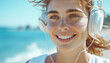 Cheerful young woman close-up portrait with freckles enjoying music on headphones with beach background. Bright smile and blue eyes, radiating joy and relaxation concept portrays leisure and happiness