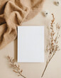 Blank paper sheet card with mockup copy space and dry floral branch and blanket cloth on neutral beige background. Minimal aesthetic wedding invitation template. Flat lay, top view