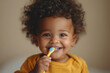 Toddler with curly hair brushes teeth with a yellow toothbrush, eyes sparkling with joy.