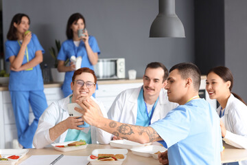 Team of doctors taking selfie while having lunch at table in hospital kitchen