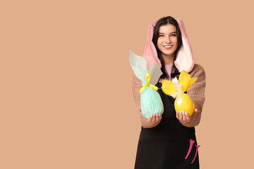 Poster - Happy female hairdresser in bunny ears with chocolate Easter eggs and hairpins on brown background