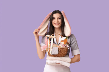 Wall Mural - Happy female chambermaid in bunny ears with Easter basket and stack of towels on purple background