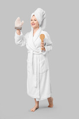 Beautiful young happy woman in bathrobe with bath mitten and massage brush waving hand on grey background