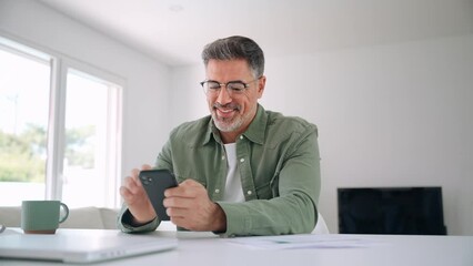 Wall Mural - Happy middle aged mature man senior entrepreneur wearing glasses holding cellphone using mobile cell phone looking at smartphone technology making banking payment sitting at home table.